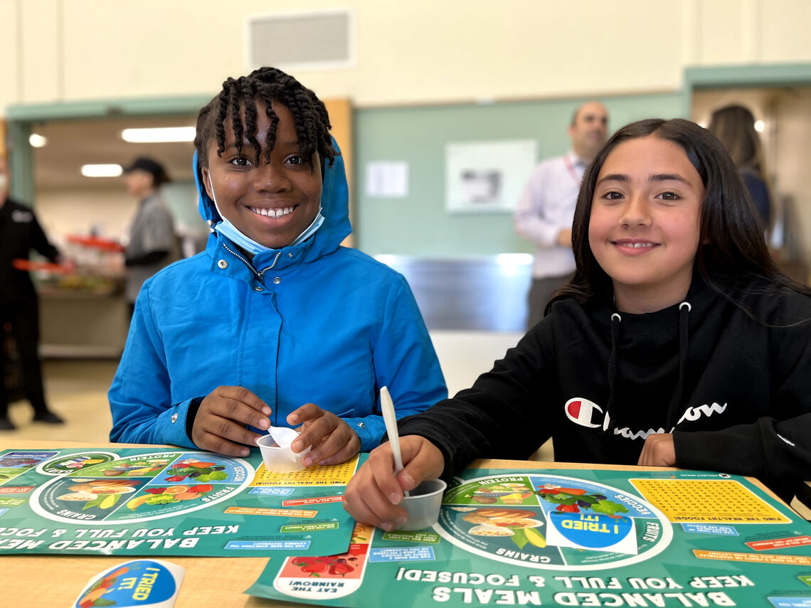 Two young girls sit at the cafeteria table enjoying a school lunch together