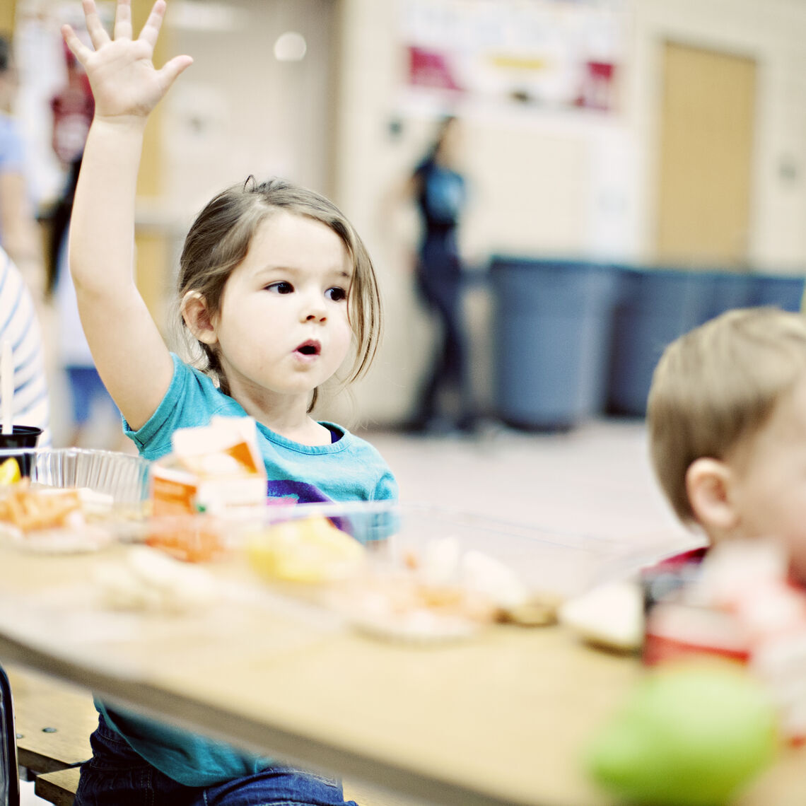 Girl eagerly raises hand at her desk in a classroom. She has breakfast on her desk.